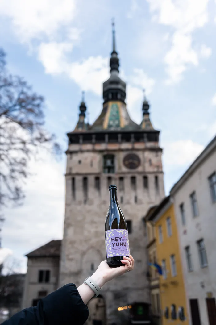 O mână ridicând o sticlă de Voyage Kombucha în fața Turnului cu Ceas din Sighișoara