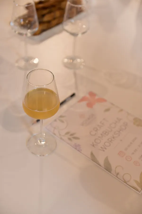 Un pahar de kombucha lângă ghidul tipărit Craft Kombucha Workshop, pe o masă albă